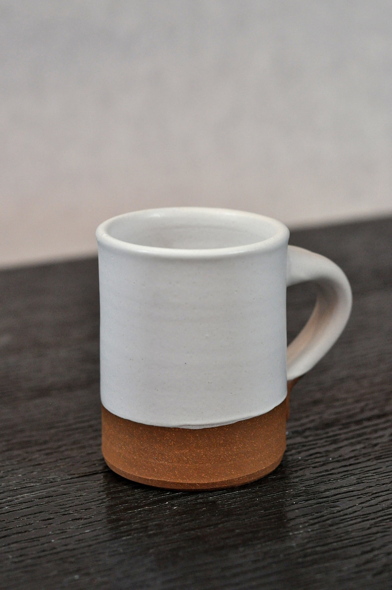 May include: A white ceramic mug with a brown clay base. The mug has a rounded handle.
