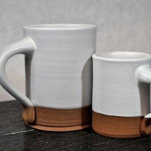 May include: Two ceramic mugs, one larger than the other, with a white glaze and a brown rim. The mugs have a simple, rounded shape and a handle.