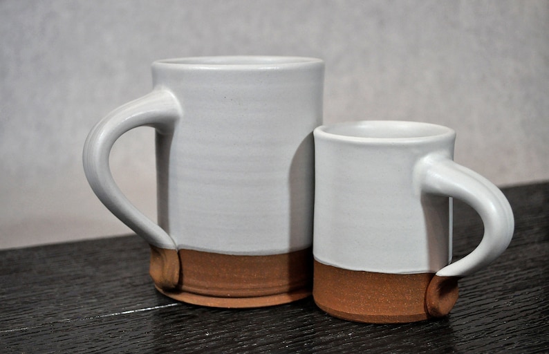 May include: Two ceramic mugs, one larger than the other, with a white glaze and a brown rim. The mugs have a simple, rounded shape and a handle.