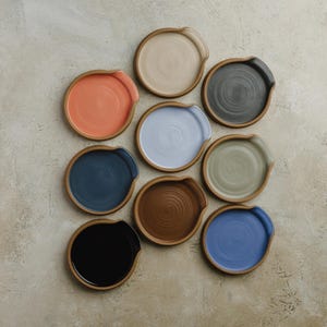 May include: A collection of nine circular ceramic spoon rests arranged in a grid. Each piece features a distinct solid color including black, dark blue, orange, light blue, beige, brown, dark gray, sage green, and bright blue with a rim.