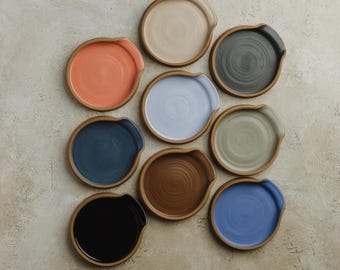 Op de afbeelding: Een verzameling van negen ronde keramische lepelhouders gerangschikt in een raster. Elk stuk heeft een eigen effen kleur, waaronder zwart, donkerblauw, oranje, lichtblauw, beige, bruin, donkergrijs, saliegroen en helderblauw met een rand.