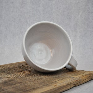 Puede incluir: Una taza de cer&aacute;mica blanca con forma redondeada y asa. La taza est&aacute; sobre una pieza de madera desgastada.