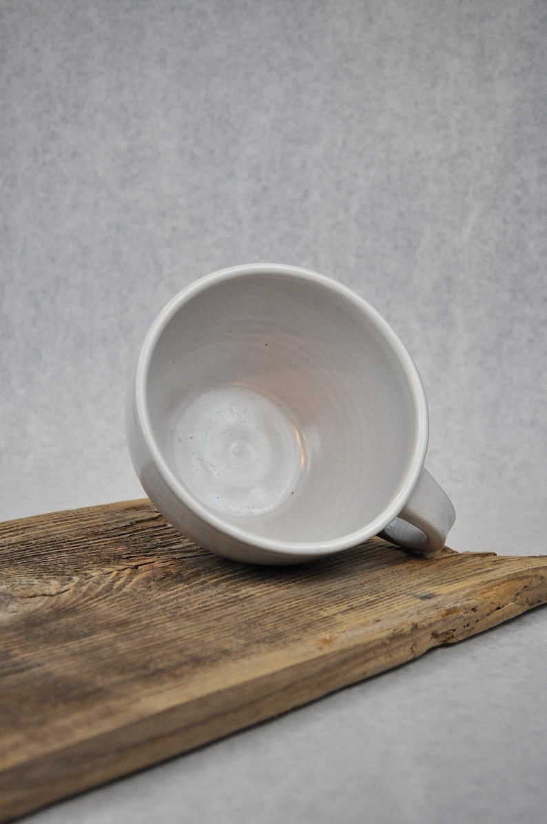 Puede incluir: Una taza de cer&aacute;mica blanca con forma redondeada y asa. La taza est&aacute; sobre una pieza de madera desgastada.