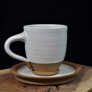 May include: A white ceramic coffee mug with a brown rim and base, sitting on a brown saucer. The mug and saucer are on a wooden surface.