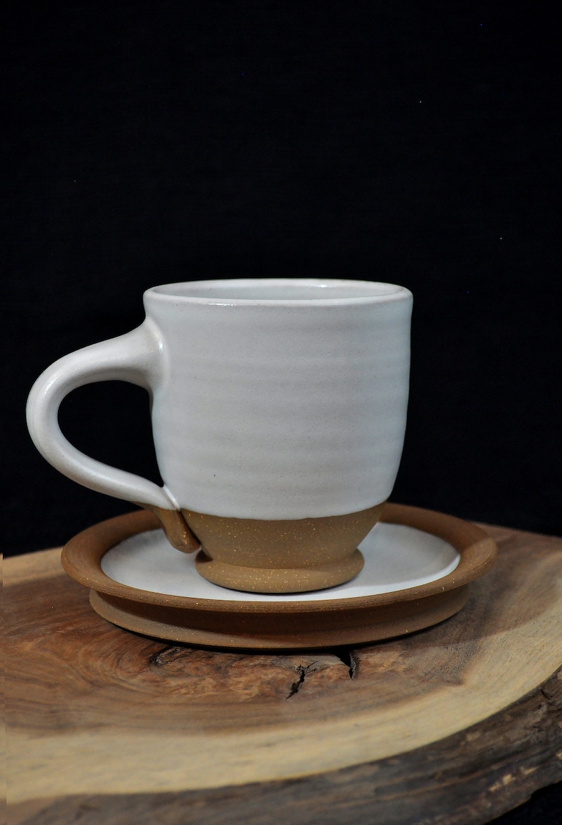 May include: A white ceramic coffee mug with a brown rim and base, sitting on a brown saucer. The mug and saucer are on a wooden surface.