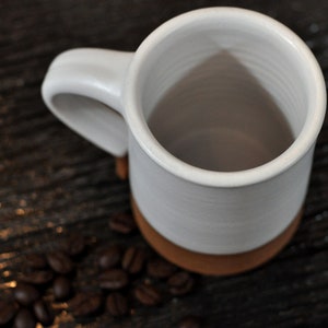 May include: White ceramic mug with a brown rim, empty and sitting on a dark wooden surface with coffee beans scattered around it.
