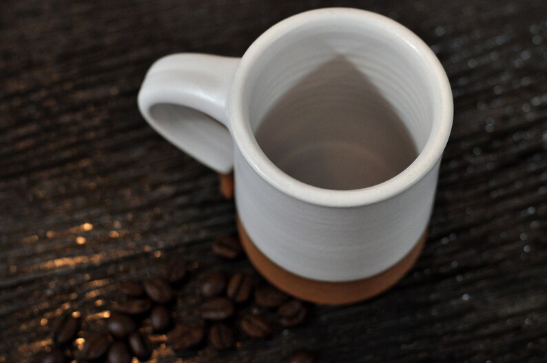 May include: White ceramic mug with a brown rim, empty and sitting on a dark wooden surface with coffee beans scattered around it.