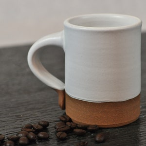 May include: A white ceramic mug with a brown rim and a handle. The mug is sitting on a dark wooden surface with coffee beans scattered around it.