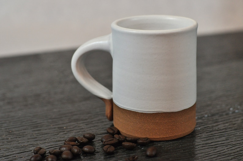 May include: A white ceramic mug with a brown rim and a handle. The mug is sitting on a dark wooden surface with coffee beans scattered around it.