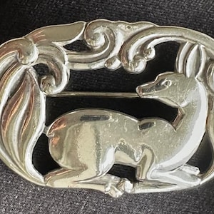 May include: Silver brooch depicting a deer in a reclining position, set within an oval frame with ornate detailing. The brooch, with a pin fastening, is crafted from a shiny metal and is suitable for a jacket or coat.