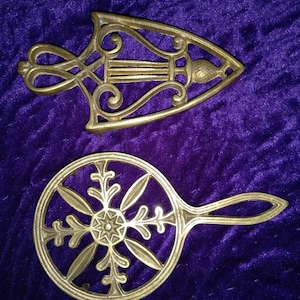 May include: Two antique brass trivets. One is shaped like a pointed oval with a decorative design. The other is round with a floral design.