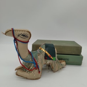 May include: A beige camel figurine with a blue and red yarn bridle and a green and gold saddle with fringe. The camel is sitting on top of two green books.