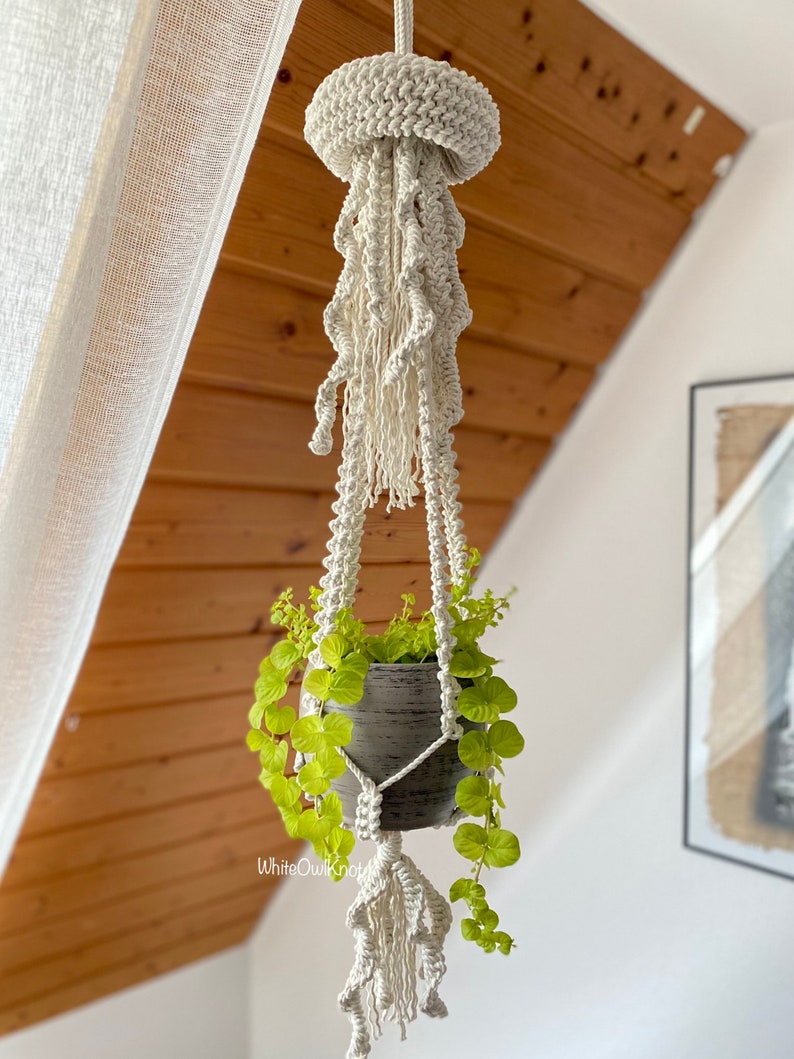 May include: A white macrame plant hanger shaped like a jellyfish with a gray pot and green plant hanging from it.
