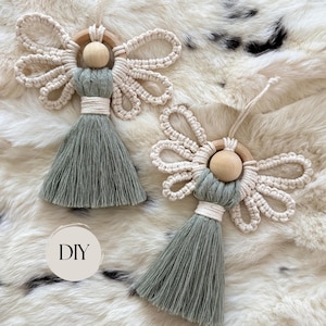 May include: Two macrame angel ornaments featuring sage green cord bodies, natural wooden bead heads, and cream-colored cord wings attached to wooden rings. Each ornament has a tassel skirt and a hanging loop. A circular tag with the text DIY is visible.