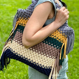 May include: A large, multi-colored macrame tote bag featuring horizontal stripes in navy blue, cream, brown, dark green, and mustard yellow. The bag includes long fabric fringe along the sides and a strap adorned with smooth, round wooden beads.
