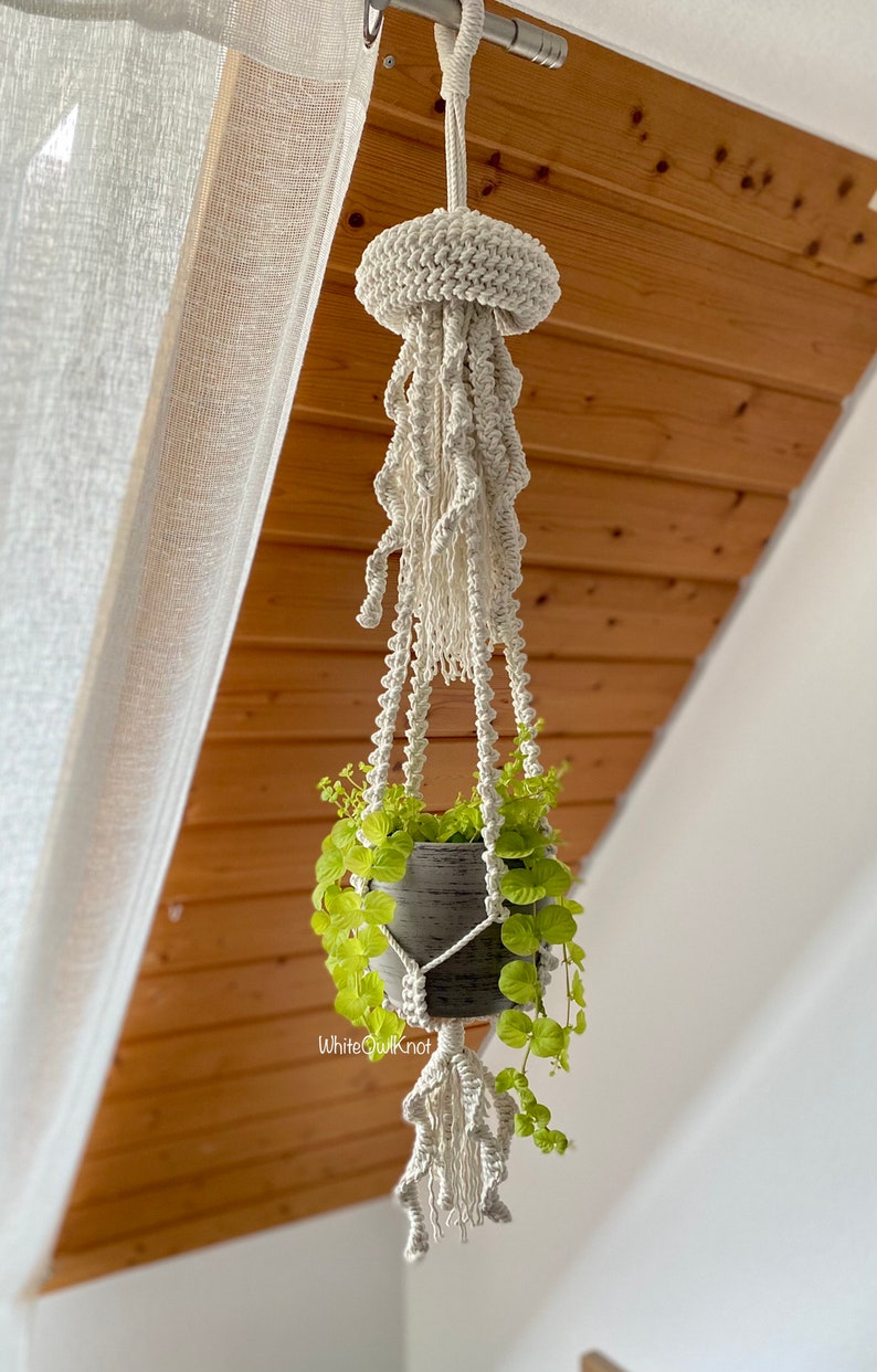 May include: A white macrame plant hanger shaped like a jellyfish with a green plant in a gray pot hanging from a ceiling.