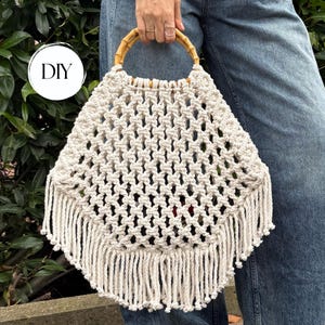 May include: A cream-coloured macrame handbag featuring a diamond-patterned knotted design, a circular bamboo handle, and a long fringe trim along the bottom edge. The bag includes a circular white graphic label with the black text DIY.