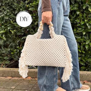 Op de afbeelding: Een off-white macramé handtas met een geweven ruitpatroon, lange franjes aan beide zijkanten en twee handvatten versierd met kleine houten kralen. Een cirkelvormige witte afbeelding met de zwarte tekst DIY is zichtbaar in de linkerbovenhoek.