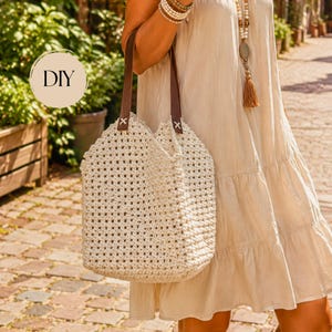 Puede incluir: Un bolso de ganchillo color crema con un patrón de rejilla cuadrada y dos correas de cuero marrón oscuro sujetas con costuras en cruz blancas. El bolso tiene una abertura superior y una base rectangular, con el texto DIY visible en un círculo.