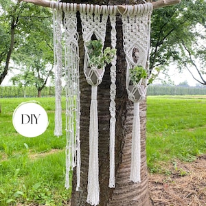 May include: A white macrame wall hanging suspended from a wooden branch, featuring intricate knotted patterns and two integrated plant holders containing green ivy plants. The design includes long fringe tassels and a circular white DIY label.