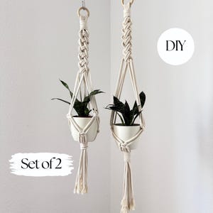May include: Two macrame plant hangers with white ceramic pots, each holding a green plant. The hangers are made of cream-colored rope and feature wooden rings at the top. The text "DIY" and "Set of 2" are also visible.