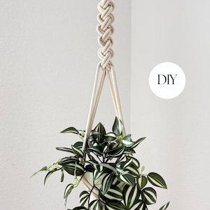 Op de afbeelding: Een crèmekleurige macramé plantenbak, met een witte pot met een groen en wit gestreepte plant. Het woord "DIY" staat in een witte cirkel rechtsboven. De plantenbak hangt tegen een witte muur.