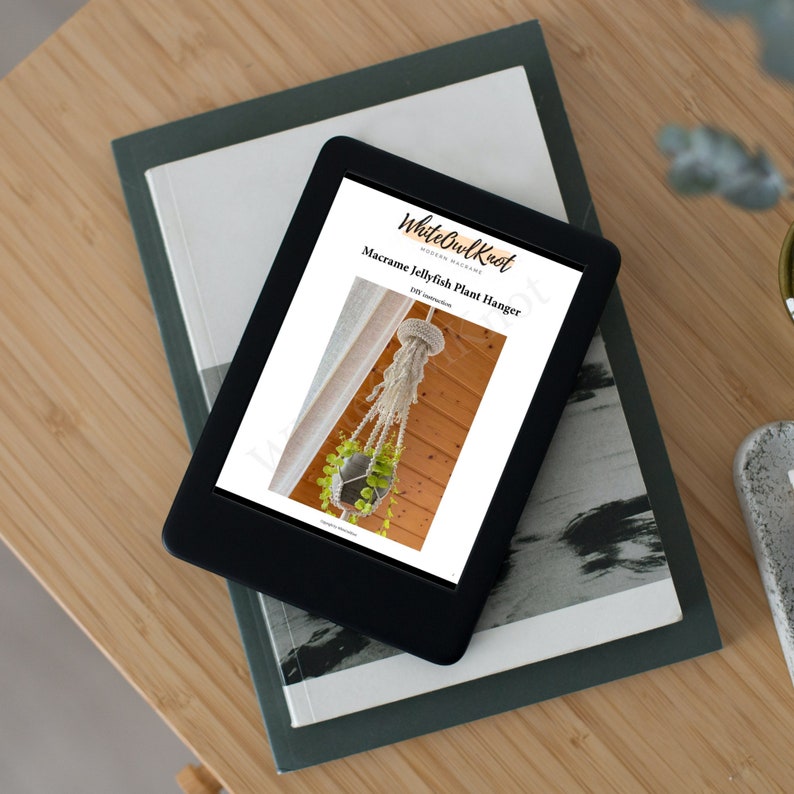 May include: A digital download for a macrame jellyfish plant hanger DIY project. The image shows a white macrame plant hanger with a green plant in a pot hanging from a wooden ceiling. The text on the page reads "White Owl Knot" and "Macrame Jellyfish Plant Hanger".