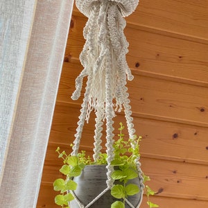 May include: A white macrame plant hanger shaped like a jellyfish with a gray pot and green plant hanging from it.