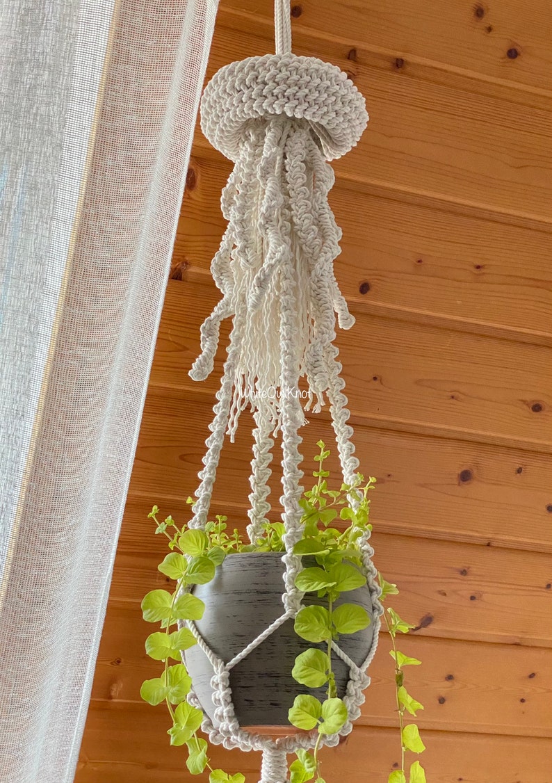 May include: A white macrame plant hanger shaped like a jellyfish with a gray pot and green plant hanging from it.