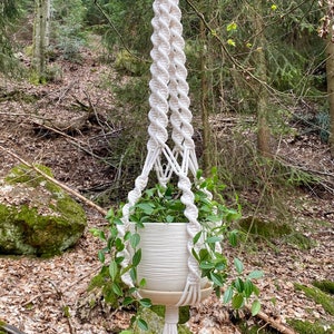 May include: A white macrame plant hanger with a white ceramic pot and green plant hanging from a tree branch in a forest setting.