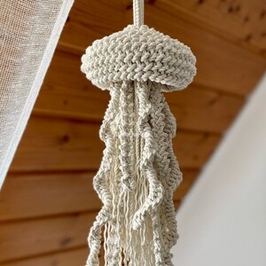May include: A white macrame plant hanger shaped like a jellyfish, hanging from a ceiling hook.