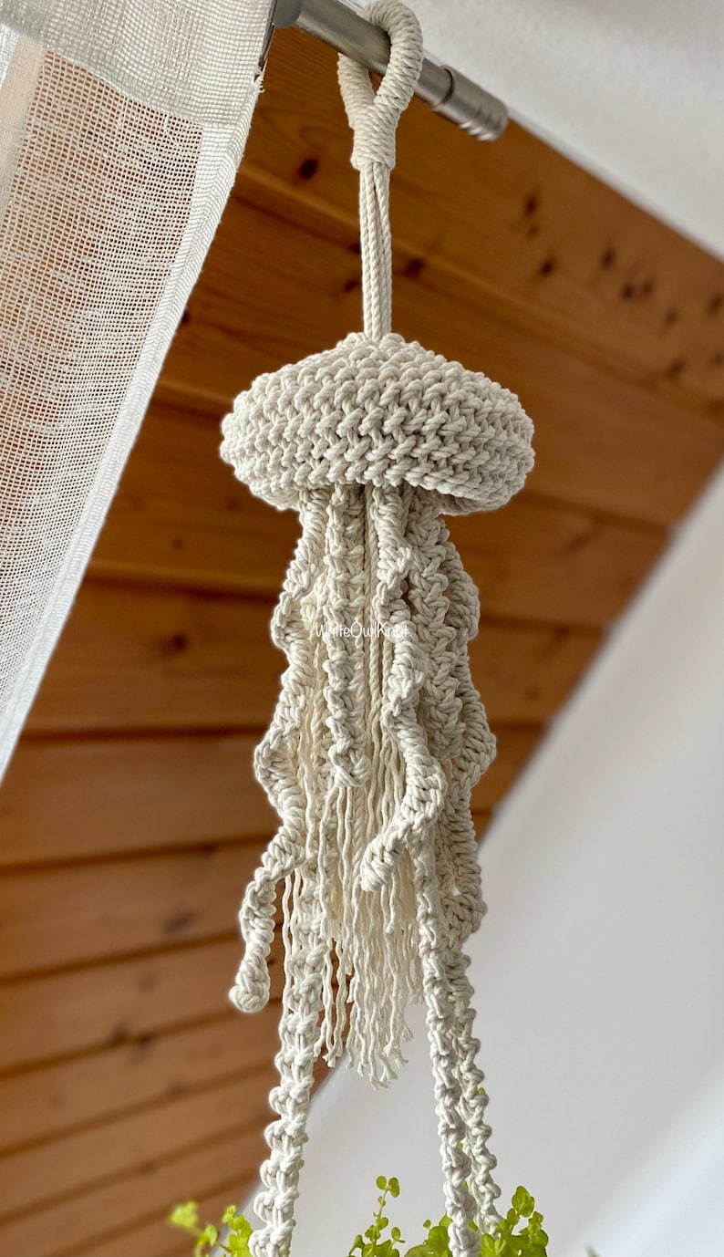 May include: A white macrame plant hanger shaped like a jellyfish, hanging from a ceiling hook.