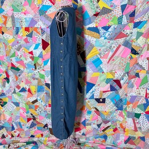 May include: A sleeveless, button-down denim dress in a medium blue wash. The dress has a V-neck and a long, straight shape. It features small, round buttons down the front and a slit at the hem. The dress is displayed on a white dress form.