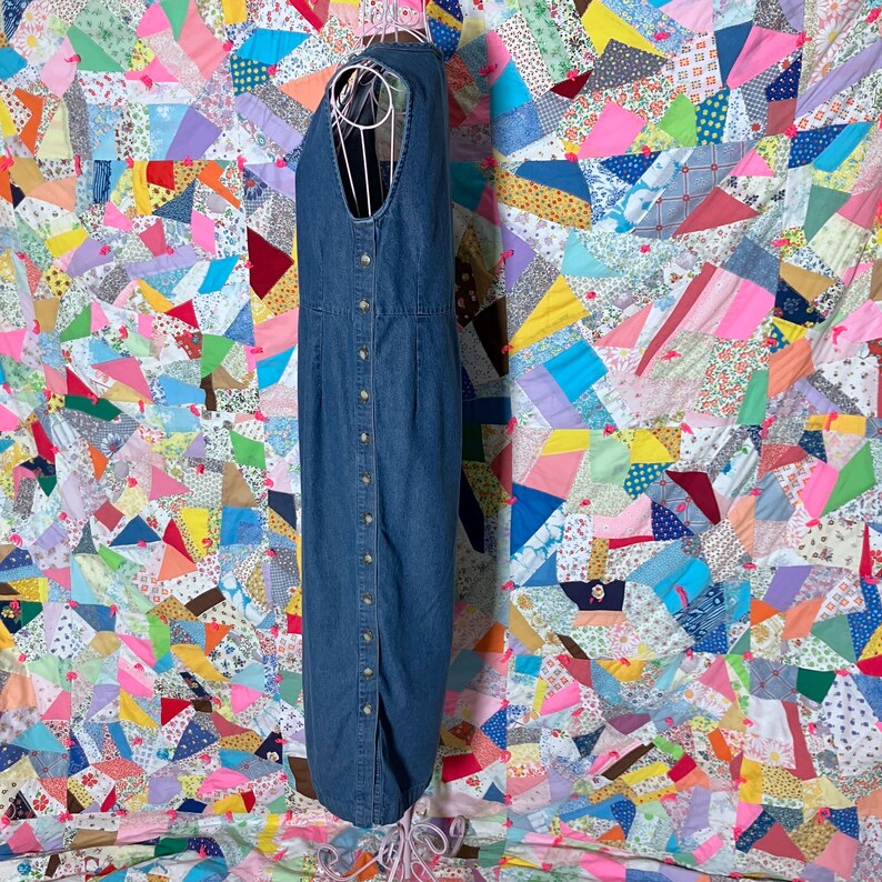 May include: A sleeveless, button-down denim dress in a medium blue wash. The dress has a V-neck and a long, straight shape. It features small, round buttons down the front and a slit at the hem. The dress is displayed on a white dress form.