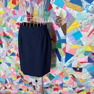 May include: A navy blue pencil skirt displayed on a pink wire dress form. The skirt has a high waist and a simple, classic design. The background is a colorful patchwork quilt with various fabric patterns.