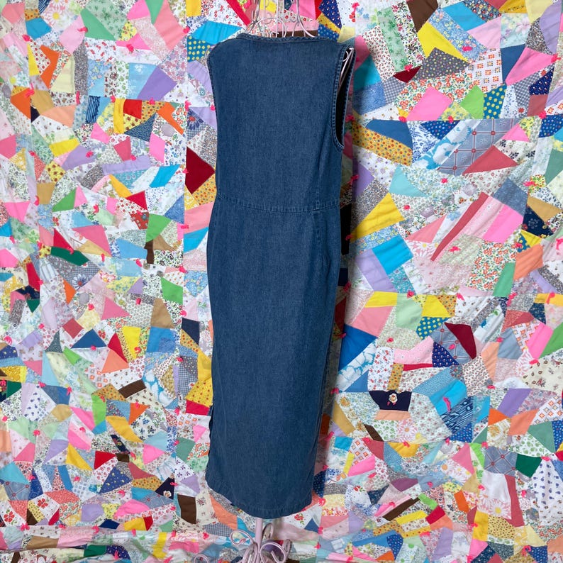 May include: A sleeveless, dark blue denim dress. The dress features a V-neck, a fitted waist, and a knee-length hem. The dress is displayed on a hanger against a colourful patchwork quilt background.