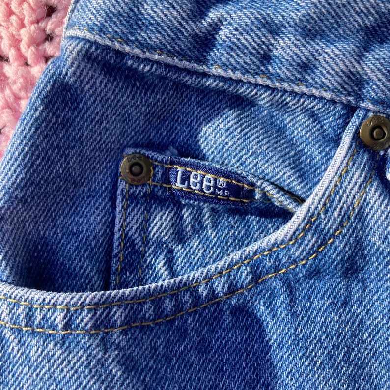 Puede incluir: Primer plano de un bolsillo de mezclilla azul con una etiqueta de la marca Lee&reg; y un remache de metal. El bolsillo tiene costuras amarillas.