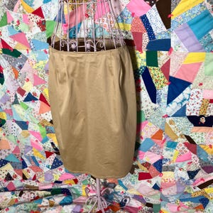 May include: A tan pencil skirt made of a smooth fabric. The skirt is on a mannequin and is displayed against a colorful patchwork quilt.