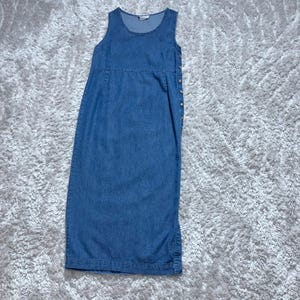 May include: A sleeveless, blue denim dress with a round neckline and a button-up side closure. The dress has a fitted bodice and a straight skirt, suitable for casual wear.