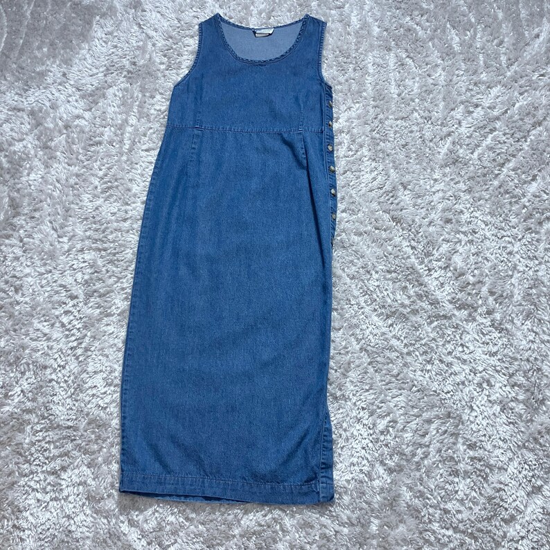 May include: A sleeveless, blue denim dress with a round neckline and a button-up side closure. The dress has a fitted bodice and a straight skirt, suitable for casual wear.