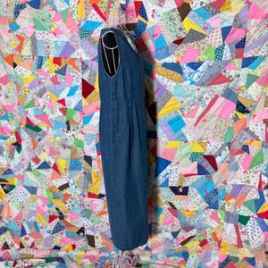 May include: A sleeveless, denim blue dress with a scoop neckline. The dress is displayed on a wire dress form. The background is a colourful patchwork quilt with various fabric patterns and colours.