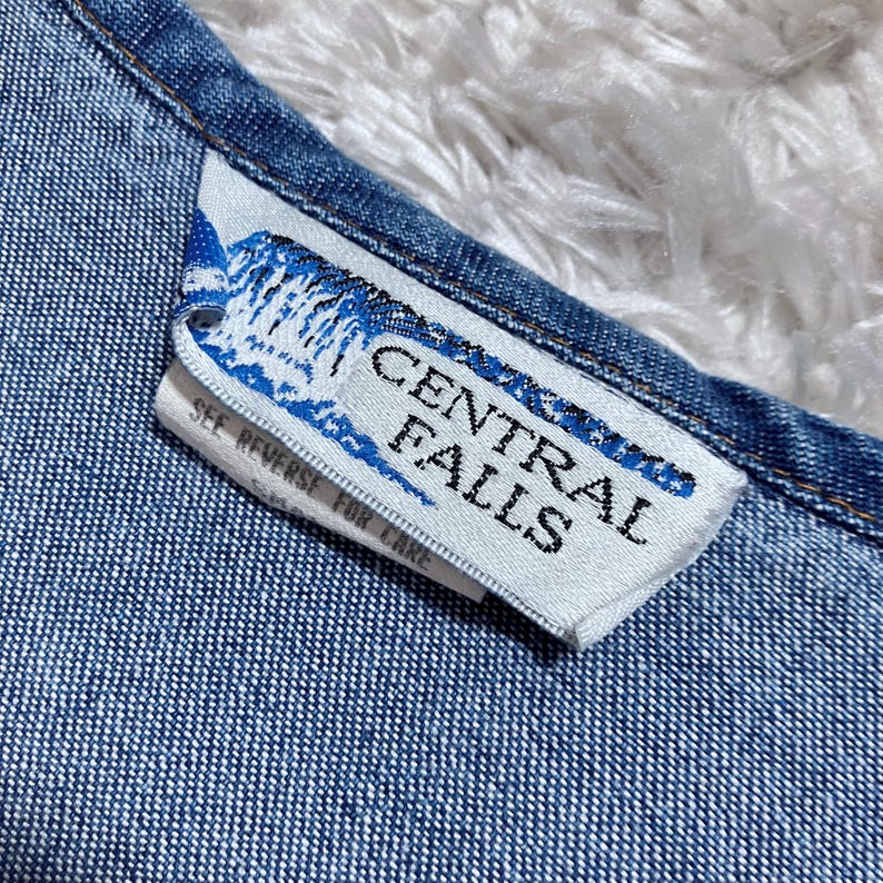 May include: Close-up of a denim garment with a white tag. The tag features the text "CENTRAL FALLS" in a bold, sans-serif font, with a blue and white graphic. The tag also includes the text "SEE REVERSE FOR CARE".