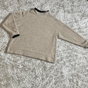 May include: A tan fleece pullover sweater with a mock neck and long sleeves. The collar and cuffs are trimmed with black fabric. The sweater is lying on a white and gray textured surface.
