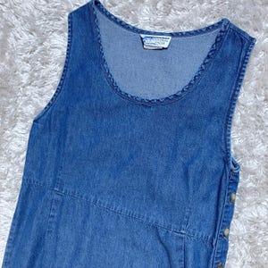 May include: A sleeveless, blue denim dress with a scoop neckline. The dress has a button closure on the side and a small tag with text. The dress has a classic, casual style.