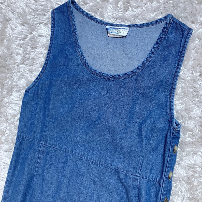 May include: A sleeveless, blue denim dress with a scoop neckline. The dress has a button closure on the side and a small tag with text. The dress has a classic, casual style.
