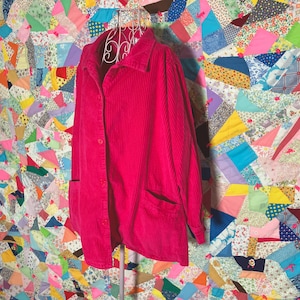 May include: A pink corduroy button-up shirt with long sleeves and two pockets. The shirt is on a white mannequin and is in front of a colorful patchwork quilt.