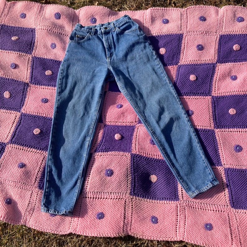 Puede incluir: Un par de pantalones vaqueros azules de mezclilla con cintura alta y pierna recta. Los vaqueros est&aacute;n colocados sobre una manta de crochet rosa y morada.
