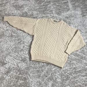 May include: A cream-colored cable-knit sweater with a crew neck. The sweater features a textured pattern of vertical and horizontal cable designs. The sleeves are long and the bottom hem is ribbed. The sweater is lying on a textured, light gray surface.