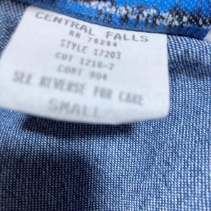 May include: A close-up shot of a white label with black text, set against a blue denim background. The label displays text including "CENTRAL FALLS", "STYLE 17203", and "SMALL".