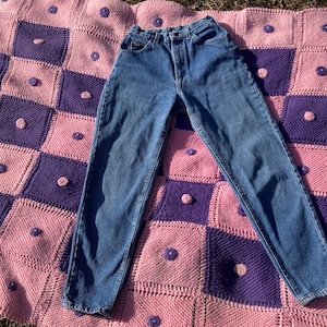 Puede incluir: Un par de pantalones vaqueros azules de mezclilla con cintura alta y pierna recta. Los pantalones vaqueros est&aacute;n colocados sobre una manta de crochet rosa y morada.